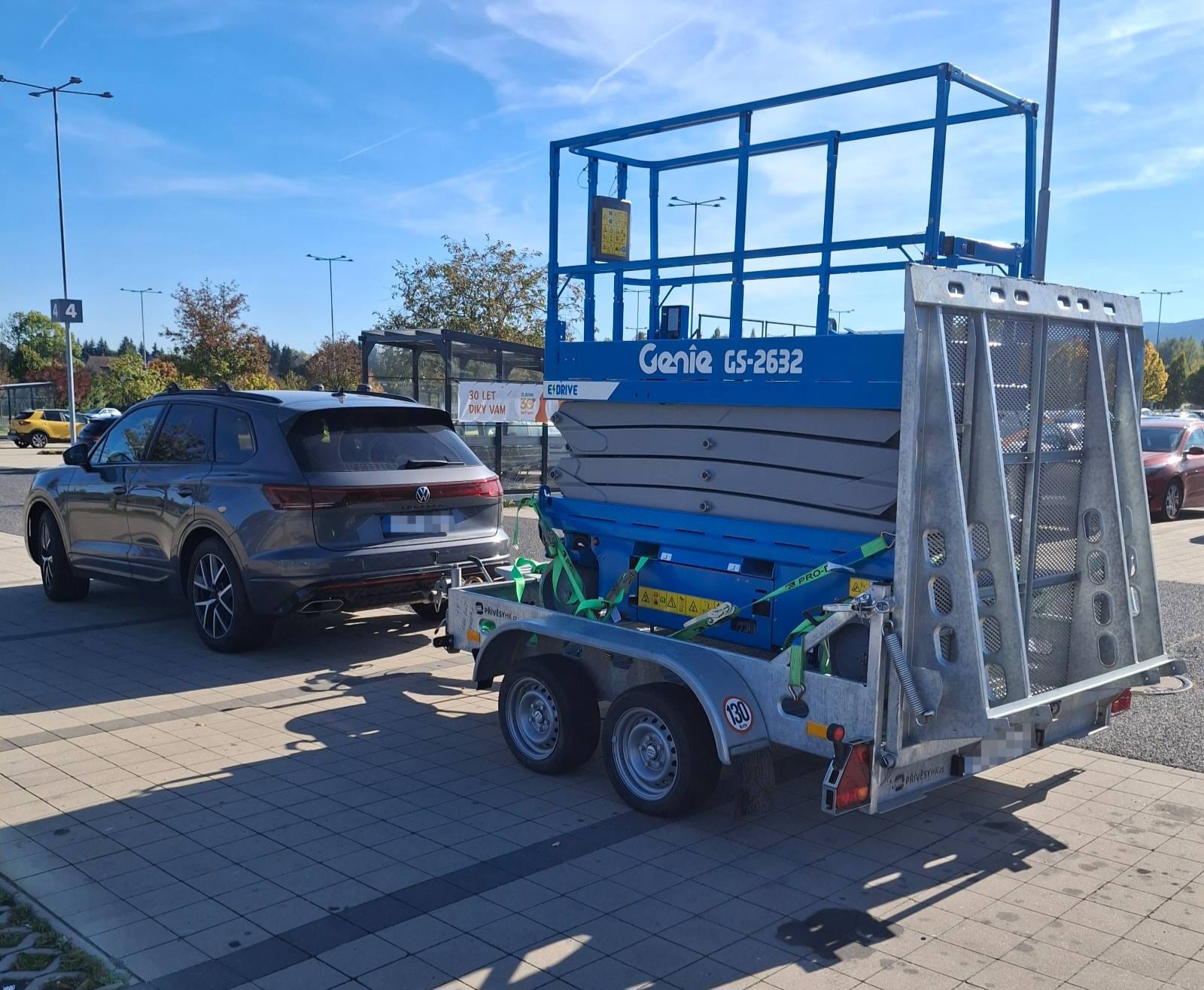 'Modrý hydraulický nakládací přívěs CS-2632 na parkovišti, vedle osobních aut, pod modrou oblohou.